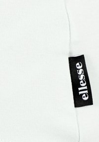 Weißer Baumwollstoff mit gerippter Textur, versehen mit einem schwarzen Etikett, auf dem das "ellesse"-Logo seitlich aufgenäht ist.