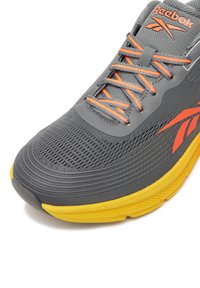 Grauer Laufschuh mit Mesh-Obermaterial, orangen Akzenten an den Schnürsenkeln und dem Logo sowie einer auffälligen gelben Gummisohle. Verfügt über strukturierte Überzüge und filigrane Nähte.