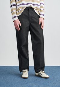 Crne, široke traperice izrađene od trapera, s čistim dizajnom i blago sjajnom teksturom, u kombinaciji s bijelim tenisicama s crnim prugama.