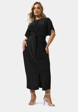 Femme aux longs cheveux blonds portant une robe midi noire à devant torsadé et des sandales argentées à talons hauts, debout sur un fond clair.