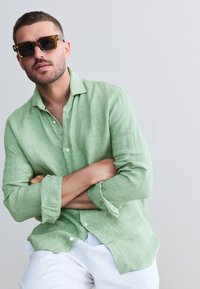 Chemise en lin vert clair, coupe décontractée, boutonnée à l'avant, avec manches retroussées, associée à des lunettes de soleil marron surdimensionnées.