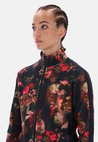 Žena s vrkočmi, oblečená v čiernej bunde so zipsom s viacfarebným kvetinovým vzorom, pozerá doľava na jednoduchom pozadí.