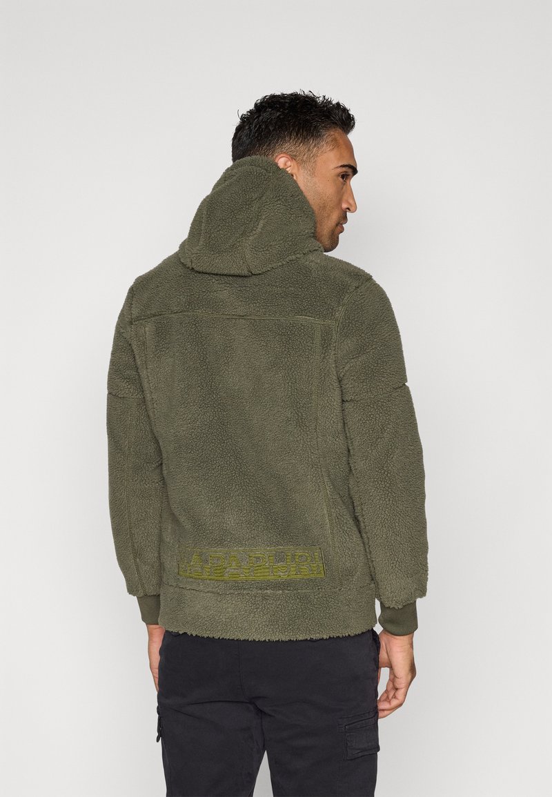 Mann, der von hinten mit einer olivgrünen Fleece-Hoodie mit hochgezogenem Kapuze und schwarzen Cargo-Hosen vor einem einfachen weißen Hintergrund steht.