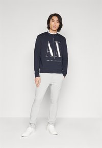 Felpa da uomo blu navy con logo "AX", collo rotondo e polsini a costine, abbinata a pantaloni slim-fit grigio chiaro e sneakers bianche.