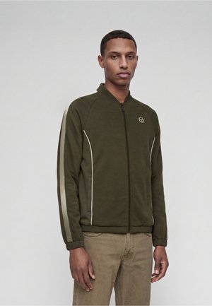 Homme portant une veste zippée vert olive avec des bandes claires sur les manches et un pantalon beige, debout devant un fond uni clair.
