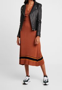 Hnědé žebrované šaty s V-výstřihem a detaily knoflíků, doplněné černou koženou motorkářskou bundou. Bílé a zelené tenisky dotvářejí vzhled.