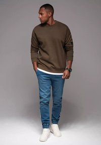 Oliwkowy sweatshirt z ściągaczami, w zestawie z jasnoniebieskimi spodniami dresowymi z bocznymi paskami i elastycznymi kostkami oraz białymi sneakersami.