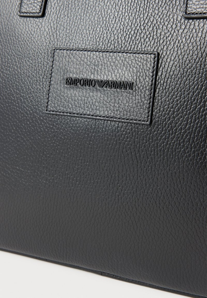 Borsa tote in pelle nera con finitura testurizzata, caratterizzata da un logo Emporio Armani impresso in rilievo su una targhetta rettangolare. Forma quadrata, doppi manici.