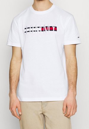 Hvid bomulds-T-shirt med broderet "TOMMY JEANS"-tekst og vandrette røde og marineblå striber tværs over brystet. Korte ærmer, rund hals.