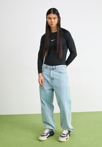 Sort langærmet top med Nike-logo, lyseblå løse jeans og sorte/grå sneakers med beige accenter på et grønt gulv.
