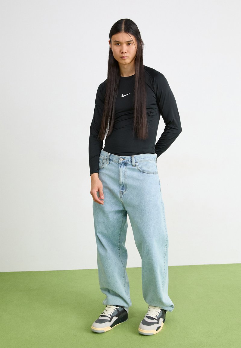 Maglietta nera a maniche lunghe con logo Nike, jeans larghi azzurri, e scarpe nere/grigie con dettagli beige su un pavimento verde.