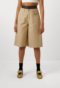 Beige Denim-Shorts mit hoher Taille und einem schlangenartigen Bund, ausgestattet mit Seitentaschen und geradem Bein, kombiniert mit goldenen Sneakers.