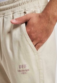 Beige cotton trousers with an elastic waistband, side pocket, and embroidered logo detail on the leg. Smooth texture and relaxed fit.