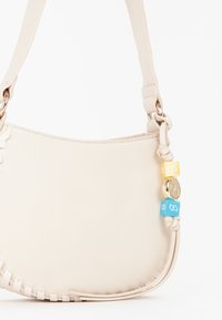 Sac à épaule en cuir couleur crème avec une forme courbée. Présente des accents tressés et des perles décoratives en jaune et bleu sur la bandoulière.