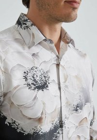 Chemise blanche avec imprimé floral noir et gris, dotée d'un col pointu et de boutons transparents, fabriquée en tissu léger avec une texture lisse.