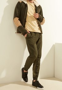 Chaqueta verde oliva con mangas de microfibra de ante en color beige, camisa beige, pantalones verde oliva y zapatillas negras contra un fondo claro.