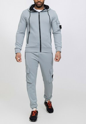 Survêtement en polaire gris avec une fermeture éclair à capuche, des poignets côtelés et des poches cargo. Accents noirs sur les zips et les chaussures avec des lacets orange.