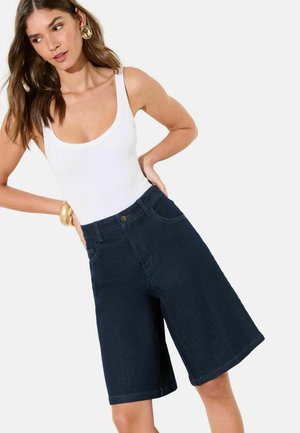 Frau in weißem ärmellosem Oberteil und dunkelblauen, kniehohen Jeansshorts, posiert mit Händen in die Hüften gestützt vor schlichtem Hintergrund.