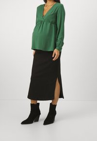 Gravid kvinna i grön långärmad blus, svart midikjol med sidospalt, svarta klackstövlar och lager av gyllene halsband.