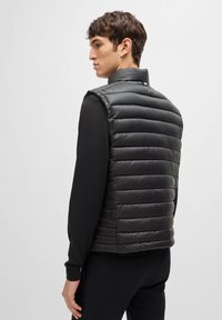 Black padded vest with a high collar, featuring a quilted design and sleek, smooth texture. Worn over a long-sleeve black shirt.