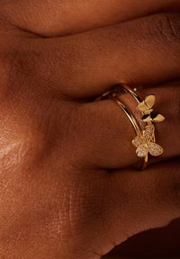 Gouden vlinder ringen, waarvan de ene met heldere stenen en een ontwerp met drie bloemblaadjes, en de andere met een solide afwerking, gepresenteerd op een gestructureerd huidoppervlak.
