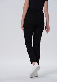 Zwarte skinny jeans met elastische tailleband, twee achterzakken en een tapse pijpdesign, gecombineerd met witte sneakers op een effen achtergrond.