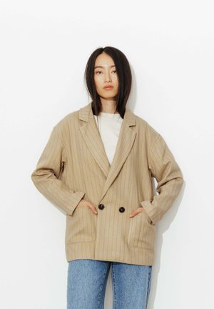 Femme aux cheveux noirs et raides porte un blazer oversize beige à fines rayures, croisé, avec des boutons noirs, par-dessus une chemise blanche et un jean bleu.