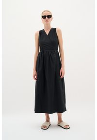 Schwarzes ärmelloses Wickelkleid mit gerafftem Taillenbund und bodenlanger Rock. Das Model trägt schwarze Sonnenbrillen und beigere Slipper.