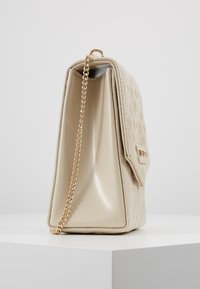 Borsa a tracolla in ecopelle beige trapuntata con manico a catena. Finitura lucida, design piegato e un fermaglio decorativo sul risvolto anteriore.