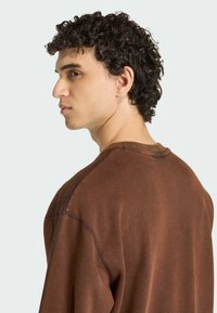 Hnedý oversized sveter s okrúhlym výstrihom, viditeľným šitím a jemne textúrovaným povrchom. Model je zobrazený v bočnom pohľade.