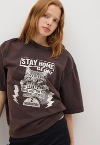 Camiseta oversized marrón con un diseño gráfico que presenta un gato y texto. Manga corta, cuello redondo y corte relajado, hecha de una tela suave.