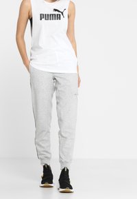 Witte mouwloze tanktop met zwart "PUMA"-logo, gecombineerd met lichtgrijze joggingsbroek met zijaccentstripen en enkels boven. Zwarte sneakers.