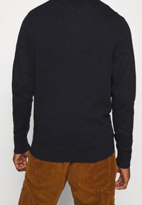Pull bleu marine foncé en tissu tricoté avec manches longues et ourlet arrondi. Associé à un pantalon en velours côtelé marron avec poches visibles.