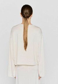 Cremefarbene, oversized, langärmelige Bluse mit verkürztem Saum. Weicher Stoff und lockere Passform. Langes Haar zu einem tiefen Pferdeschwanz frisiert.