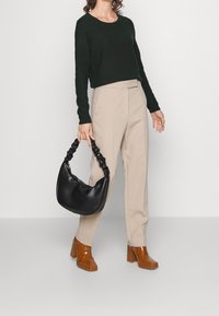 Borsa a tracolla in pelle nera con manico intrecciato. Il modello indossa un maglione a coste verde scuro e pantaloni beige su misura, abbinati a stivali lucidi marroni.