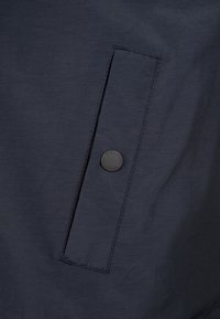 Marinblå jacktyg med en subtil textur; har en rektangulär bröstficka med en rund svart knapplogotyp.