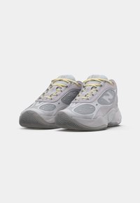 Grå og hvide sneakers i mesh og ruskind, afrundet form, teksturerede såler, gule snørrebånd og synlige logo-detaljer.