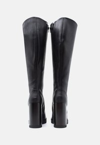 Botas altas de cuero negro con una textura smooth, punta cuadrada y tacones altos y gruesos. Cuentan con una cremallera trasera para facilitara su uso.