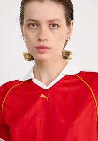 Red sports jersey with vertical stripes, white collar, and gold Puma logo. Features short sleeves with yellow accents on the shoulder seams.
