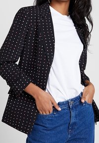 Black blazer with white and red polka dots, features a classic lapel, single-button closure, and relaxed fit over a white t-shirt.