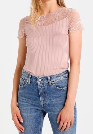 Woman wearing a pink ribbed top with lace short sleeves and high-waisted blue denim jeans, standing with hands relaxed by sides.