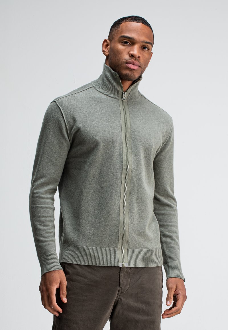 Lichtgroene ritsvest van zacht gebreid materiaal met een hoge kraag, geribde manchetten en zoom. Het heeft een rits over de volle lengte aan de voorkant.