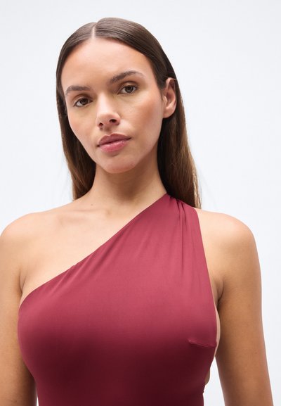 Μονόχρωμο μαγιό σε μπορντό χρώμα με ένα ώμο, λείο ύφασμα, ελάχιστη κάλυψη στη μία πλευρά και απαλό draping στο ύψος του ώμου.