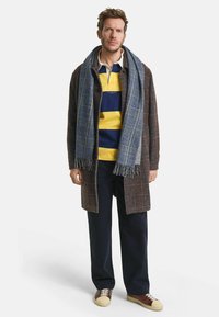 Braun karierten Mantel über einem marineblauen und gelben gestreiften Poloshirt, marineblauen Hosen und sandfarbenen Sneakers; ergänzt durch einen blauen karierten Schal mit Fransen.