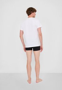 Vit kortärmad t-shirt och svarta shorts. Personen står barfota, med en neutral bakgrund och inga synliga mönster eller accenter.