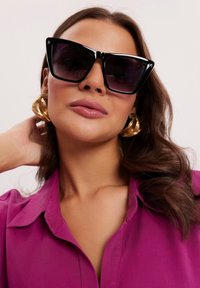 Czarne okulary w stylu kociego oka z ciemnymi szkłami, noszone z dużymi złotymi kolczykami. Osoba ma na sobie różową koszulę z kołnierzykiem, prezentując gładką skórę.