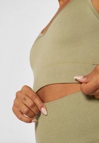 Top cropped em verde oliva com nervuras, apresentando uma banda larga com detalhe de logotipo texturizado, exibindo um tecido suave e um design ajustado. Mão posicionada na bainha.