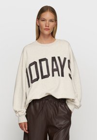 Hellgraues, oversized Sweatshirt mit "100DAYS" in dunklen Buchstaben, runder Ausschnitt und ribbelte Bündchen, kombiniert mit dunkelbraunen Lederhosen.
