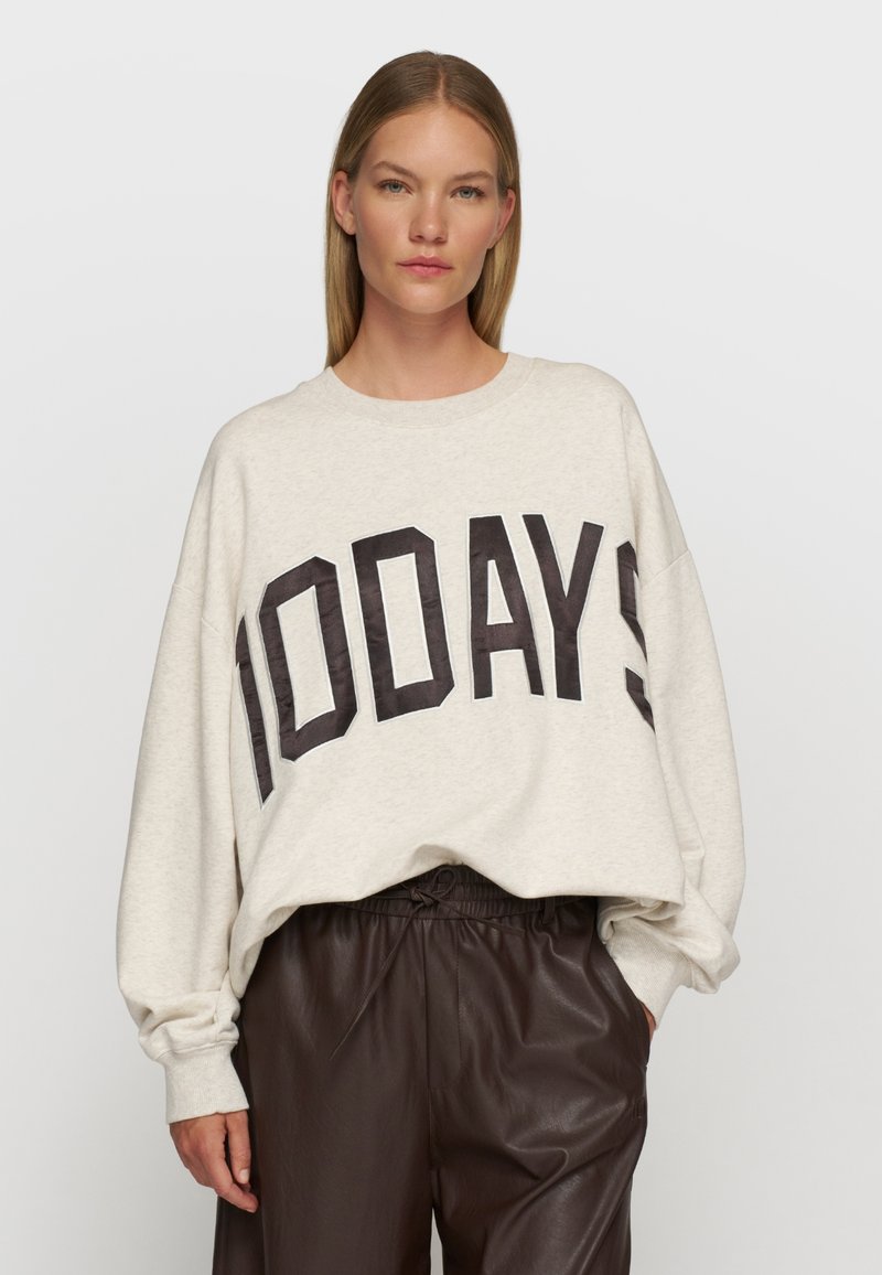 Hellgraues, oversized Sweatshirt mit "100DAYS" in dunklen Buchstaben, runder Ausschnitt und ribbelte Bündchen, kombiniert mit dunkelbraunen Lederhosen.