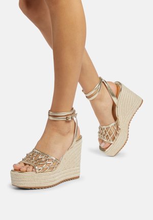 Sandales compensées avec un talon en corde tissée, des brides métalliques et un design en croix sur les orteils. La couleur est beige avec des accents métalliques.
